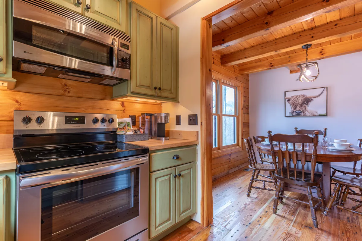 A bright kitchen scene with stainless steel appliances, green cabinets, and a wooden dining table nearby. The open setup makes it easy to prep and serve meals, and the natural lighting highlights the space’s welcoming feel.