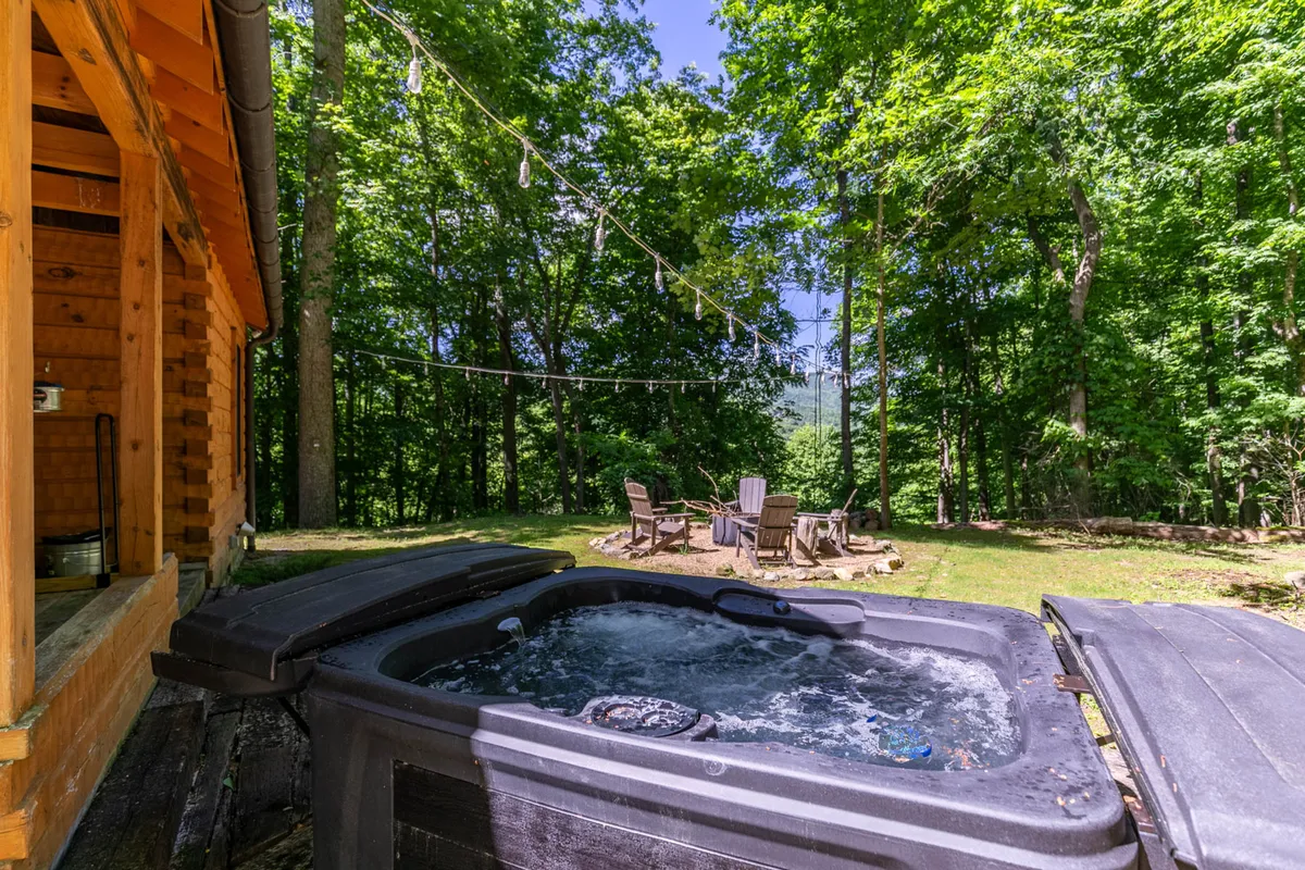 A wide exterior shot showcasing the brand-new hot tub on a wooden deck, a nearby fire pit surrounded by Adirondack chairs, and warm string lights overhead. The setting is tranquil, inviting, and clearly designed for relaxation and social gatherings under the open mountain sky.