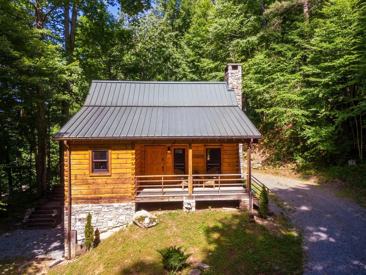 A view of the cabin’s exterior, featuring a charming wooden porch, a robust stone chimney, and surrounding trees. The porch is set up for comfortable seating, inviting guests to relax outdoors and enjoy the natural setting. The setting sun and gentle outdoor lighting create a tranquil and romantic atmosphere ideal for couples.
