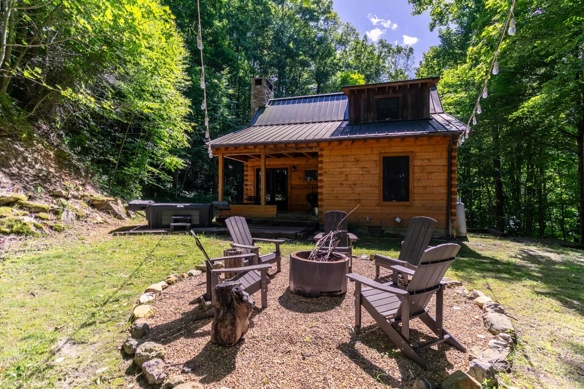 This image showcases the exterior of a log cabin with a welcoming fire pit area surrounded by Adirondack chairs and encircled by tall trees. The outdoor setting is perfect for campfires, stargazing, and family gatherings in nature.