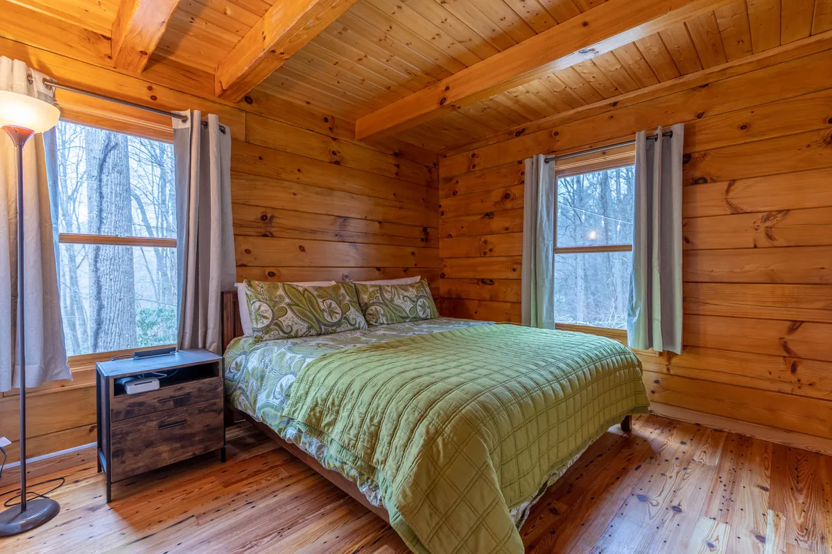 A warm and inviting bedroom featuring a cozy bed, soft lamp lighting, wooden walls, and light filtering in through curtained windows. The space feels restful and private, ideal for unwinding after a long day.