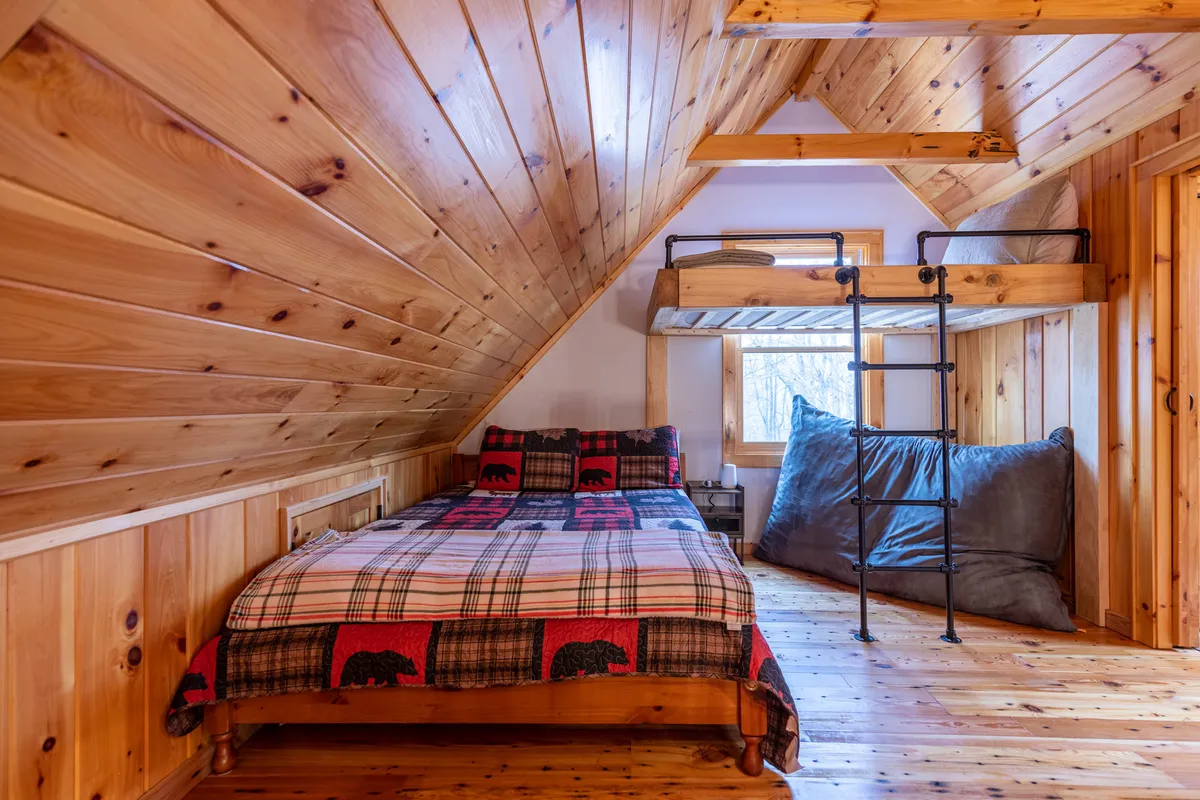 A charming bedroom with wooden walls, a bunk bed dressed in plaid bedding, large floor cushions, and ample sunlight streaming in. The room exudes warmth and is perfect for families or friends sharing a space.
