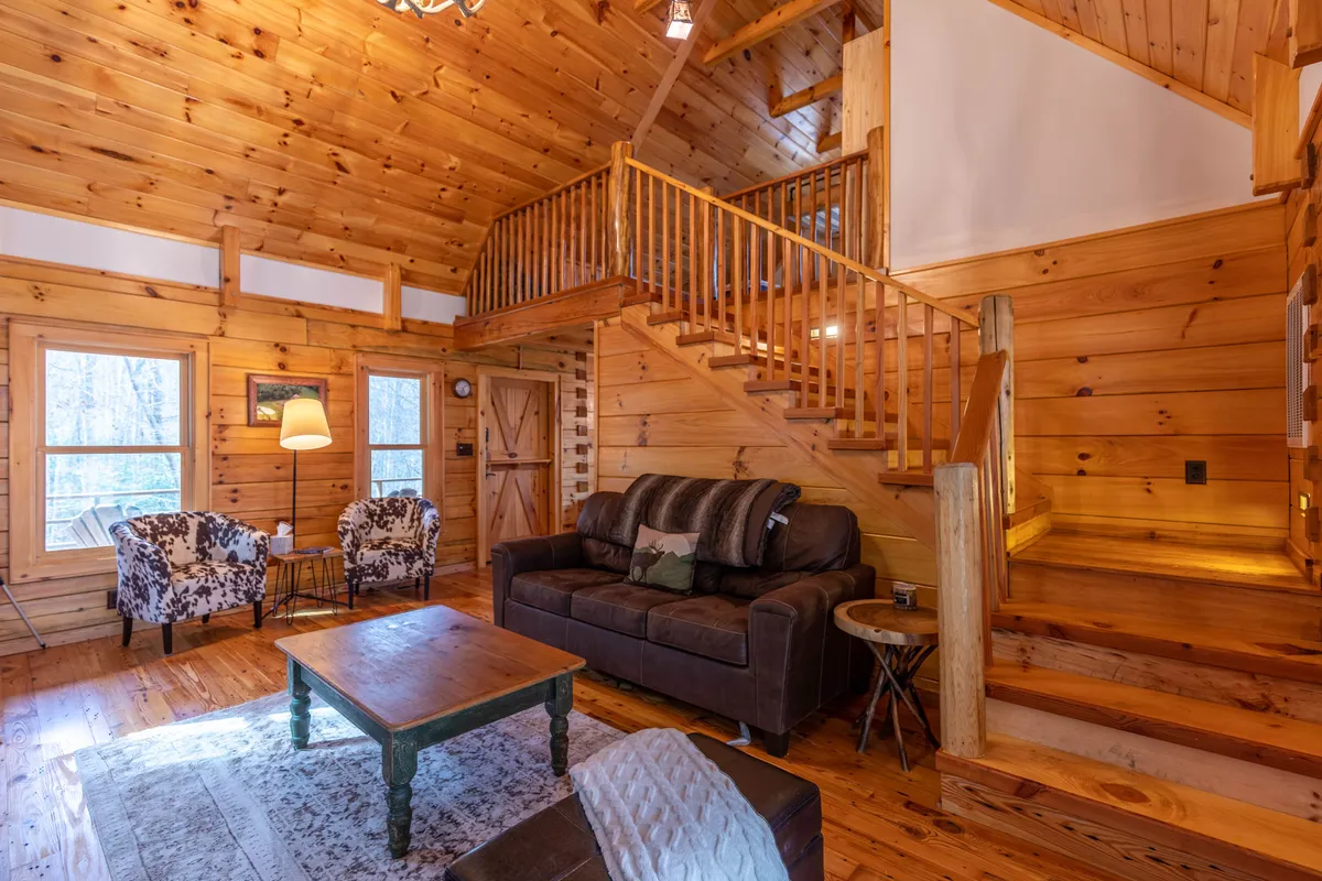 This image displays a cozy, open living space featuring a wooden staircase, comfortable sofa, armchairs, and a coffee table. Large windows provide views of the surrounding nature, creating an inviting atmosphere ideal for relaxing or gathering with friends.