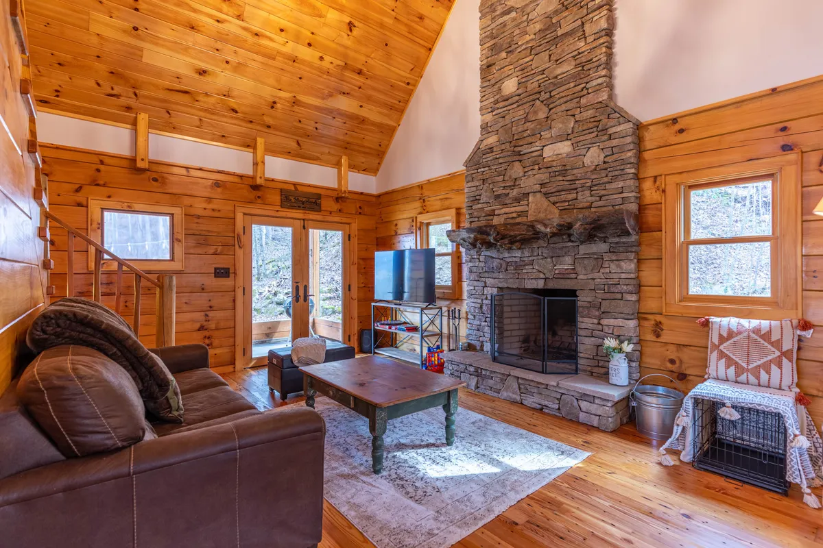 A cozy living room with a prominent stone fireplace as its focal point, surrounded by large windows that let in natural light and highlight the forest views. Comfortable sofas are arranged around the fireplace, and the wooden ceiling adds to the cabin’s rustic, welcoming atmosphere.