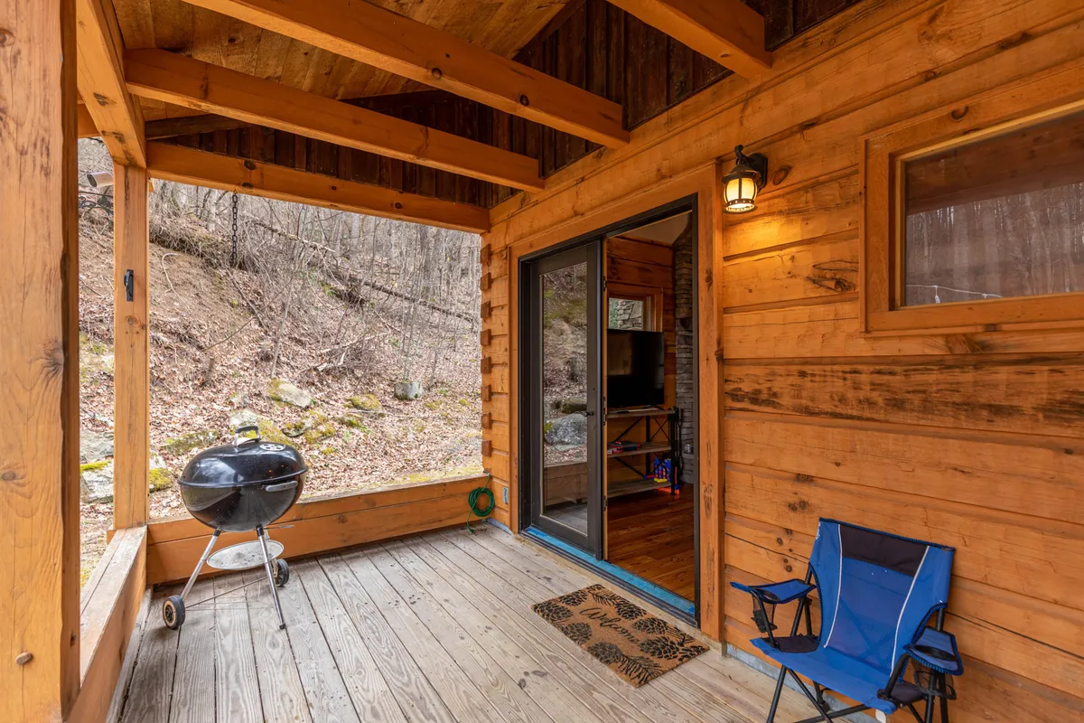This exterior image features a charming wooden structure with a grill and a blue chair set up on the porch, suggesting a relaxing outdoor space that connects directly to the surrounding nature. The doorway creates a sense of welcome and the setting evokes both adventure and comfort, making it ideal for guests looking to recharge after outdoor activities.