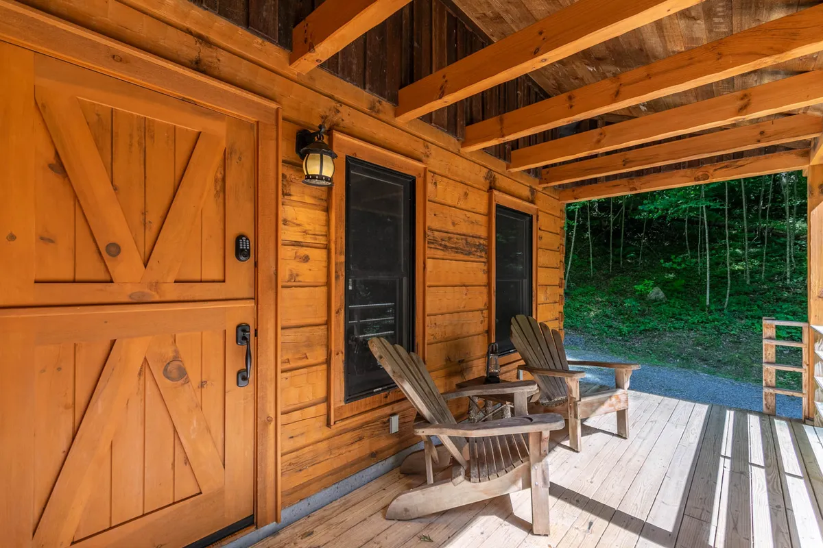 A welcoming cabin entrance framed by Adirondack chairs on the porch, set against lush natural surroundings. The warm wooden front door and subtle landscaping provide a sense of intimacy and privacy, making the space feel like a retreat for two.