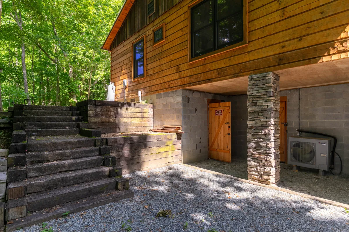 An exterior perspective highlighting the cabin’s wooden steps, stone pillars, and doorway, set against a backdrop of natural greenery. The feeling is one of arrival and anticipation, capturing the moment before an adventure begins or the satisfaction of returning to a comfortable retreat.