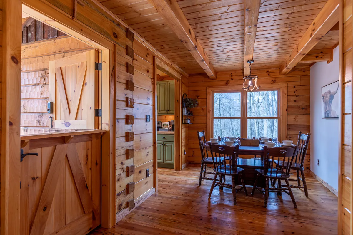 A spacious dining area featuring a sturdy wooden table under rustic beams, with large windows providing panoramic views of the surrounding forest. The bar area and door suggest easy movement between kitchen and dining spaces, reinforcing the open-concept layout.