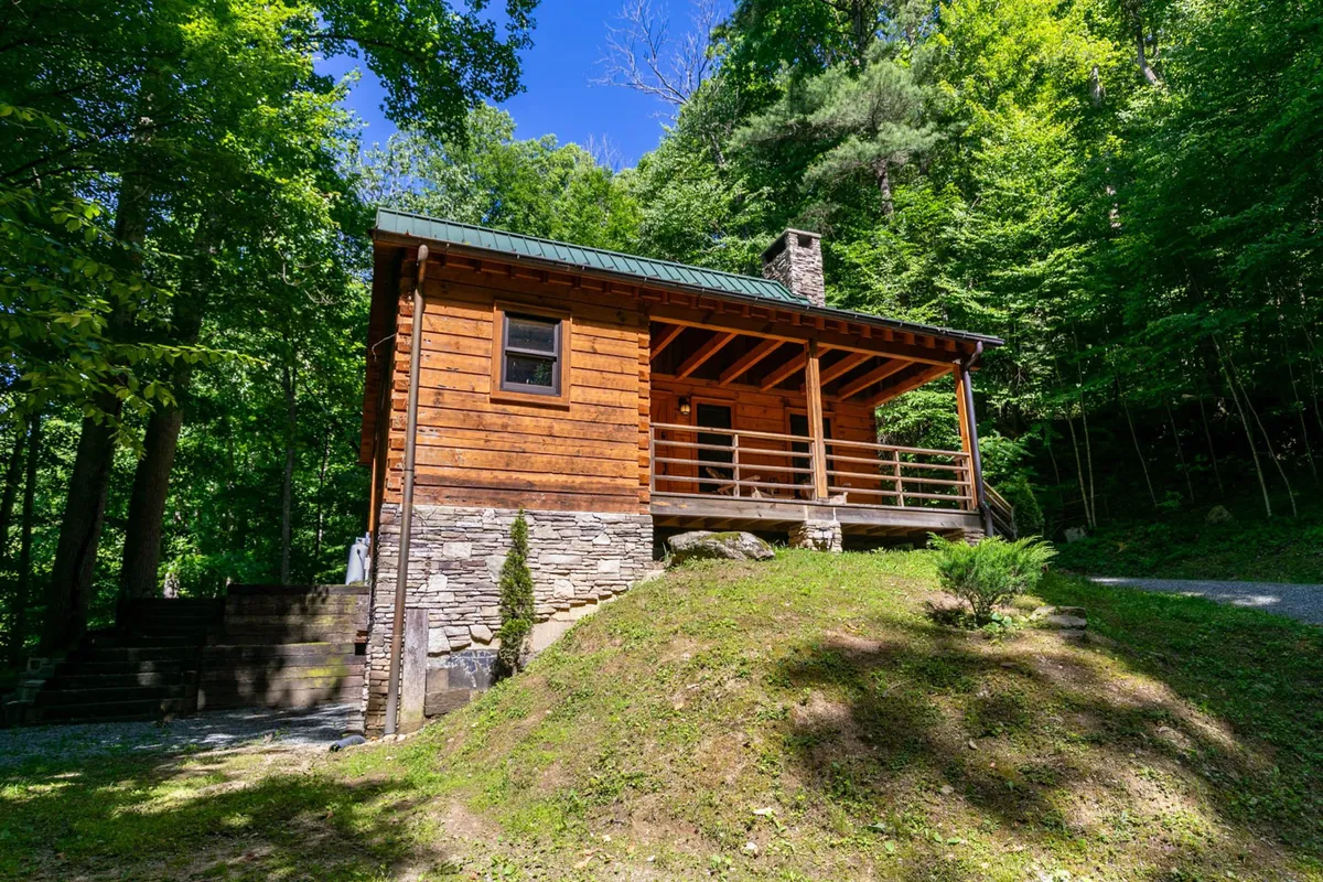 The scene presents a peaceful wooden cabin with a stone chimney, wide porch, and lush trees all around. The porch is ideal for relaxing after outdoor adventures or gathering to watch the sunset, while the natural surroundings emphasize the property's connection to nature.